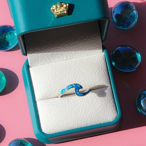 May include: A silver ring with a blue wave design, set in a teal jewelry box with a gold crown emblem. The ring is surrounded by blue glass beads on a pink surface.