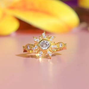 Peut inclure: Bague dor&eacute;e avec un motif de soleil, orn&eacute;e d'une pierre centrale transparente et de petites pierres. La bague pr&eacute;sente des d&eacute;tails complexes sur le bandeau, sur fond rose.