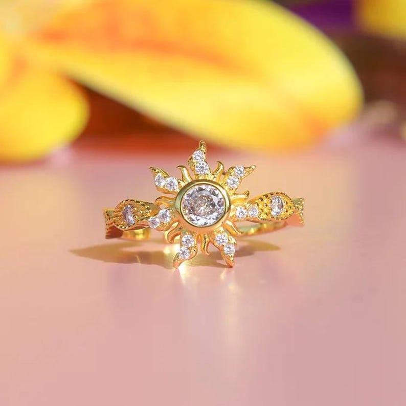 Peut inclure: Bague dor&eacute;e avec un motif de soleil, orn&eacute;e d'une pierre centrale transparente et de petites pierres. La bague pr&eacute;sente des d&eacute;tails complexes sur le bandeau, sur fond rose.
