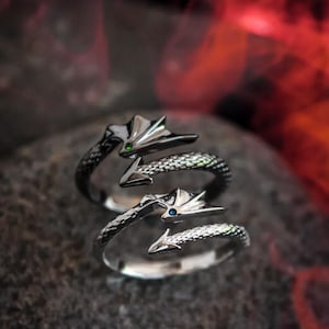 Könnte beinhalten: Zwei silberne Drachenringe mit detaillierten Schuppen und Drachenkopf-Designs. Ein Ring hat einen grünen Edelstein, der andere einen blauen Edelstein. Die Ringe werden auf einer dunklen, strukturierten Oberfläche präsentiert.