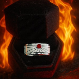 May include: A silver ring with a red gemstone center, set against a white sunburst design. The ring is displayed in a black ring box, with a fiery orange background. The ring's design is reminiscent of the Japanese flag.
