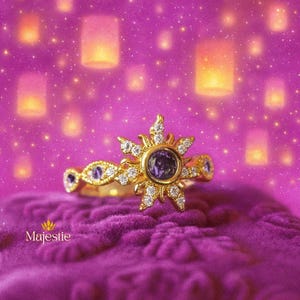 Peut inclure: Une bague en or avec un motif de soleil, orn&eacute;e d'une pierre pr&eacute;cieuse violette centrale et de diamants. La bague est pos&eacute;e sur une surface de velours violet, avec un fond de lanternes flottantes et d'&eacute;toiles. Le mot "Majestie" est visible.