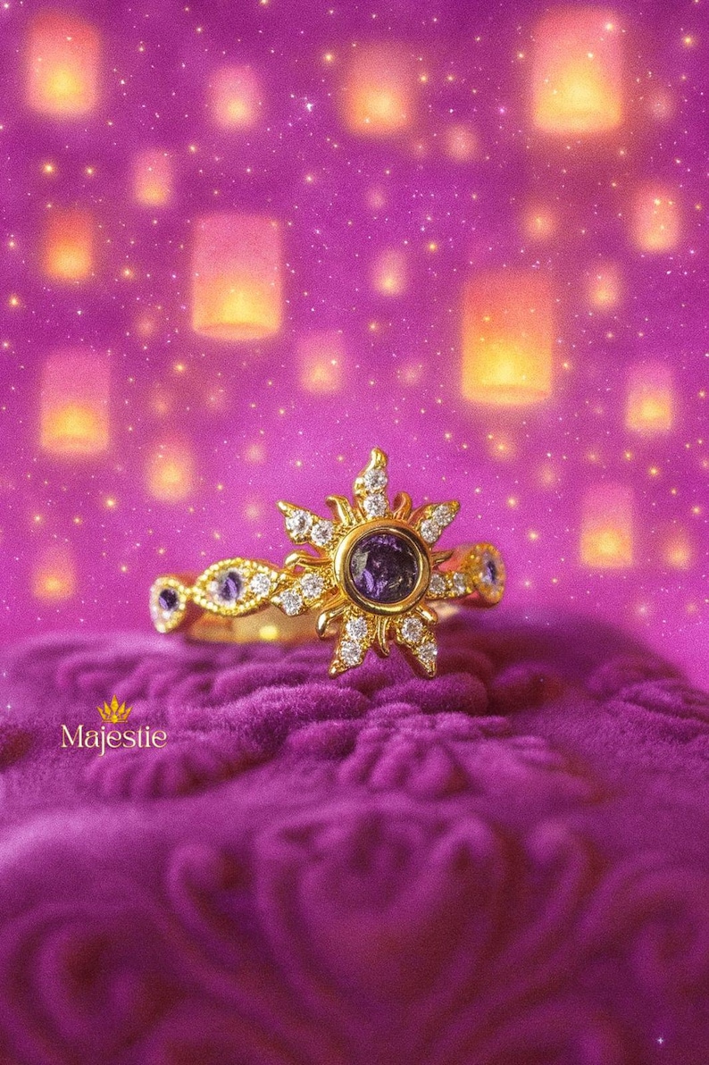 Peut inclure: Une bague en or avec un motif de soleil, orn&eacute;e d'une pierre pr&eacute;cieuse violette centrale et de diamants. La bague est pos&eacute;e sur une surface de velours violet, avec un fond de lanternes flottantes et d'&eacute;toiles. Le mot "Majestie" est visible.