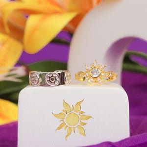 Peut inclure: Deux bagues, une argent&eacute;e et une dor&eacute;e, sont pr&eacute;sent&eacute;es sur un bloc blanc avec un motif de soleil. La bague argent&eacute;e a un motif circulaire, tandis que la bague dor&eacute;e pr&eacute;sente un motif en forme d'&eacute;clat de soleil avec une pierre centrale. Fleurs jaunes et tissu violet en arri&egrave;re-plan.