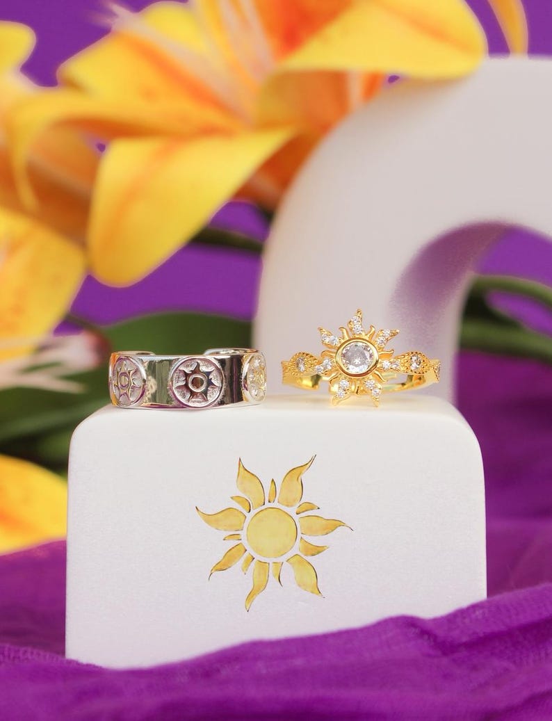Peut inclure: Deux bagues, une argent&eacute;e et une dor&eacute;e, sont pr&eacute;sent&eacute;es sur un bloc blanc avec un motif de soleil. La bague argent&eacute;e a un motif circulaire, tandis que la bague dor&eacute;e pr&eacute;sente un motif en forme d'&eacute;clat de soleil avec une pierre centrale. Fleurs jaunes et tissu violet en arri&egrave;re-plan.
