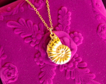 Könnte beinhalten: Eine goldfarbene Halskette mit Anhänger, die ein spiralförmiges Muscheldesign aufweist. Der Anhänger hängt an einer zarten Kette und liegt auf einer strukturierten, magenta-farbenen Oberfläche. Die Halskette ist ein Schmuckstück.