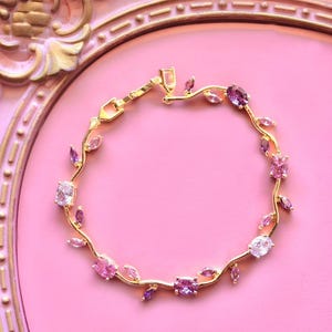 Pode incluir: Uma pulseira dourada com um design floral, com pedras preciosas rosa e roxas. A pulseira está sobre um fundo rosa, com uma moldura decorativa dourada. A pulseira é uma joia.