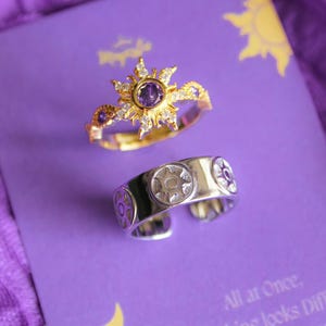 Peut inclure: Deux bagues sur une surface violette. L'une est dor&eacute;e avec un motif de soleil et une pierre centrale violette. L'autre est argent&eacute;e avec un embl&egrave;me de soleil. Le fond comprend du texte et un graphisme de soleil.
