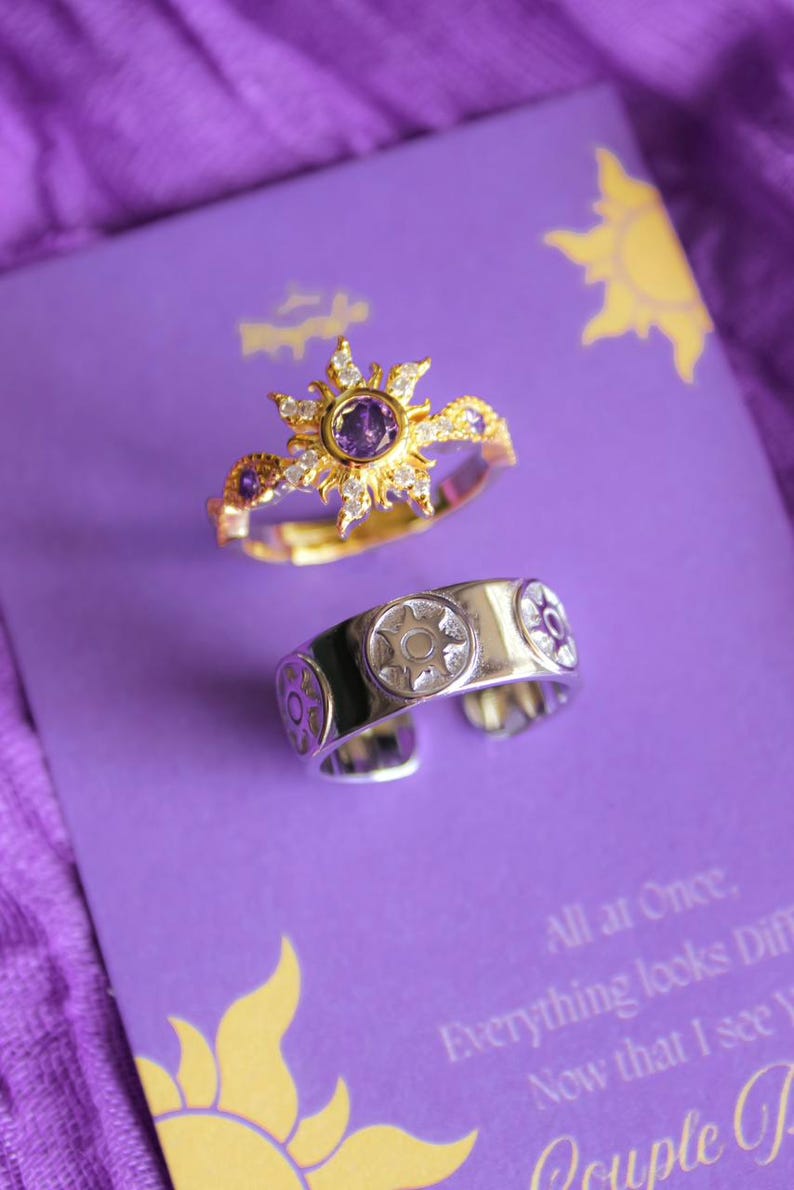 Peut inclure: Deux bagues sur une surface violette. L'une est dor&eacute;e avec un motif de soleil et une pierre centrale violette. L'autre est argent&eacute;e avec un embl&egrave;me de soleil. Le fond comprend du texte et un graphisme de soleil.