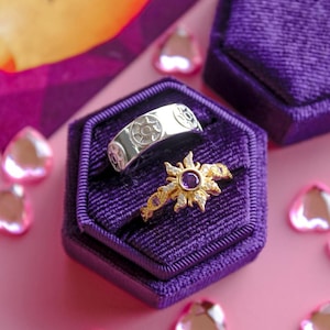 Peut inclure: Deux bagues présentées dans un écrin en velours violet. L'une est en argent avec un motif de soleil, l'autre en or avec une pierre précieuse violette centrale et des pierres blanches. Le fond est rose avec des décorations en forme de cœur.