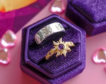 Pode incluir: Dois anéis exibidos em uma caixa de anel de veludo roxo. Um anel é prateado com um design de sol, e o outro é dourado com uma pedra preciosa roxa central e pedras brancas. O fundo é rosa com decorações em forma de coração.