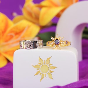 Peut inclure: Deux bagues sur un bloc blanc avec un motif de soleil. L'une est argent&eacute;e avec un motif de soleil r&eacute;p&eacute;t&eacute;. L'autre est dor&eacute;e, avec une pierre violette centrale entour&eacute;e de petites pierres et de cristaux. Fleurs jaunes et tissu violet en arri&egrave;re-plan.