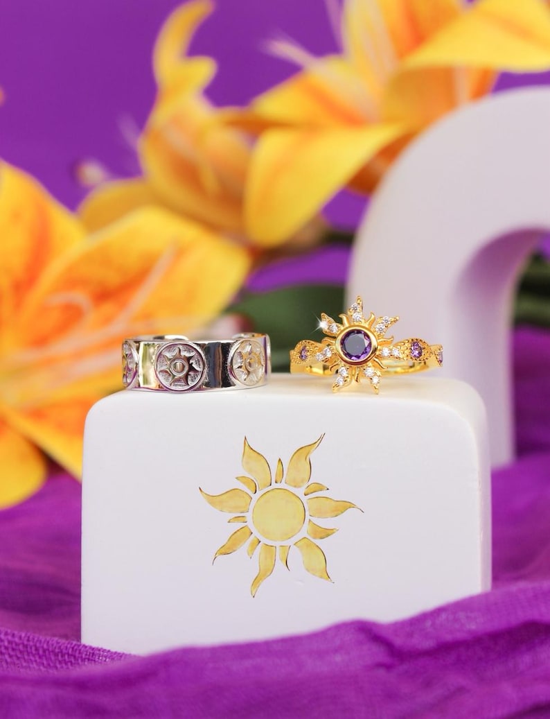 Peut inclure: Deux bagues sur un bloc blanc avec un motif de soleil. L'une est argent&eacute;e avec un motif de soleil r&eacute;p&eacute;t&eacute;. L'autre est dor&eacute;e, avec une pierre violette centrale entour&eacute;e de petites pierres et de cristaux. Fleurs jaunes et tissu violet en arri&egrave;re-plan.