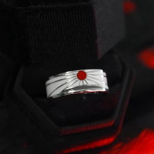 Könnte beinhalten: Ein silberner Ring mit weißem Emaille-Design, das einen roten Kreis in der Mitte zeigt, der einer aufgehenden Sonne ähnelt. Der Ring wird in einer schwarzen Samt-Ringbox mit dunklem Hintergrund präsentiert.