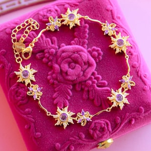 May include: A gold-colored bracelet featuring sunburst and floral charms with purple center stones. The bracelet is displayed on a pink velvet jewelry box with a rose design.
