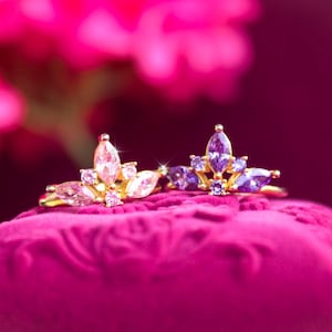 May include: Two gold-toned rings with marquise-cut gemstones in shades of pink and purple. The rings are set on a plush, magenta surface, with a blurred pink background. The gemstones are arranged in a floral or crown-like design.