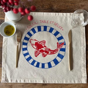 Peut inclure: Un set de table beige avec un motif de poisson rouge et bleu, une fourchette et un couteau dorés, et le texte "SOLEIL, TABLE ET DÉLICE." Un petit bol d'huile d'olive et un verre sont sur la table en bois.