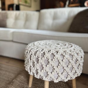 May include: A round, beige macrame ottoman with a light gray fabric seat and wooden legs. The macrame design covers the sides of the ottoman. The ottoman is placed on a brown rug in front of a white couch.