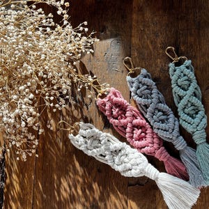 Puede incluir: Cuatro llaveros de macramé en blanco, rosa, gris y verde azulado, cada uno con un cierre dorado y una borla. Los llaveros están colocados sobre una superficie de madera junto a una ramita de flores secas.
