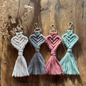 Puede incluir: Cuatro llaveros de macramé en forma de corazón en blanco, gris, rosa y turquesa. Cada llavero tiene un cierre dorado y una borla. Los llaveros están colocados sobre una superficie de madera, con flores secas en el fondo.