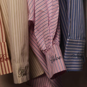 May include: Close-up of several striped shirts in various colours, including red, pink, blue, and cream. Each shirt cuff is embroidered with a name in cursive script. The shirts are hanging, showcasing the cuffs.