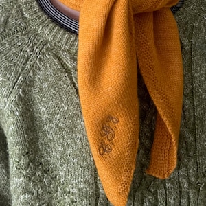 May include: Mustard yellow knitted scarf with a small embroidered design. The scarf is draped over a green cable-knit jumper with a striped collar. The scarf is made of a soft, textured material.