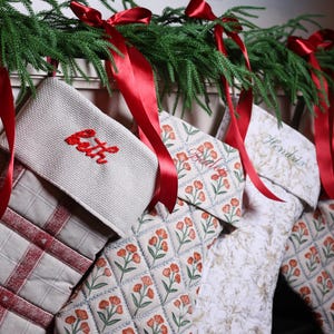 May include: A collection of Christmas stockings with personalized names embroidered in red. The stockings feature various patterns, including floral and plaid designs, and are adorned with red satin ribbons. Greenery is draped above the stockings.