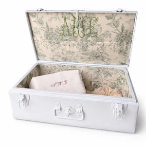 May include: White metal trunk with an open lid, revealing a floral patterned interior. The interior features a folded white cloth with embroidered initials and a small pouch. The trunk has metal latches and a handle.
