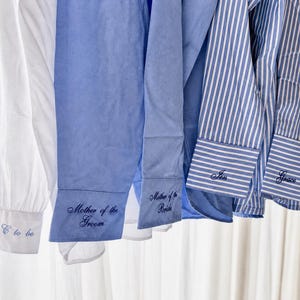 Pode incluir: Close-up de várias camisas em tons de branco e azul, algumas com riscas azuis e brancas. As punhos são bordados com nomes e títulos como "Mrs C to be", "Mother of the Groom" e "Mother of the Bride."