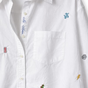 Könnte beinhalten: Weißes Button-Down-Hemd mit gestickten Aufnähern. Zu den Aufnähern gehören ein Coca-Cola-Logo, ein blaues Blumendesign, Würfel, ein Gehirn, ein Blitz und Oliven auf einem Zahnstocher. Der Schriftzug "Mi Amor" ist blau auf der Blende gestickt.