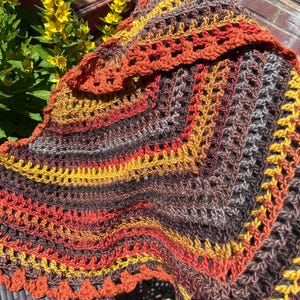 May include: A triangular crochet shawl featuring a variegated pattern of autumn-inspired colours including deep brown, burnt orange, and bright yellow. The textile displays an open, lacy stitch construction with a scalloped edge along the border.