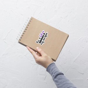 May include: A brown spiral-bound notebook with a sticker that reads "I'm a Libra, diplomacy is my weapon" in purple and black. The notebook is held at an angle against a white background.