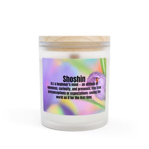 Könnte beinhalten: Eine weiße Kerze mit Holzdeckel. Das Etikett zeigt das Wort "Shoshin" mit einer Definition und einem lila Blumenbild. Die Kerze soll Achtsamkeit und eine Anfängermentalität fördern.