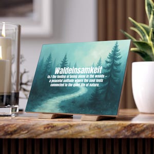 Könnte beinhalten: Dekoratives Holzschild mit dem Wort "Waldeinsamkeit" und seiner Definition in weißem Text, vor einer türkisfarbenen Waldszene. Das Schild steht auf einem Holzständer, mit einer Kerze und einer Pflanze im Hintergrund.