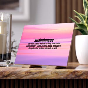 May include: A wooden block featuring the word "Suaimhneas" in black, with a definition beneath. The background displays a gradient of pink, purple, and yellow. The block is supported by two small wooden stands.
