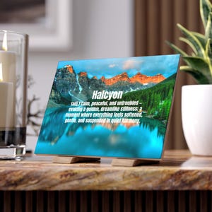 May include: A wooden block displaying the word "Halcyon" and its definition, set against a mountain and lake scene. The image features a vibrant landscape with a blue lake, green trees, and a blue sky. The block sits on a wooden stand.
