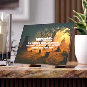 May include: A decorative wooden block featuring the word "Taphophile" with its definition, set against a cemetery scene with a sunset. The block is supported by a wooden stand.
