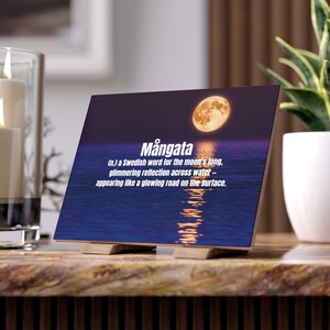 May include: A wooden block featuring the word "Mångata" and its definition, set against a moonlit water scene. The image shows the moon's reflection. The block is supported by two small wooden stands.