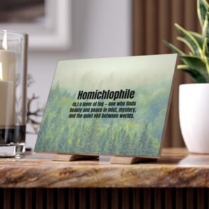 May include: A wooden sign displaying the word "Homichlophile" and its definition. The image shows a misty forest. The sign is supported by a small wooden stand.