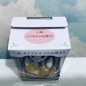 Pu&ograve; includere: Una scatola bianca con testo giapponese e un'etichetta ovale rosa. L'etichetta recita "ニ-98 メイプルネコのお母さん". All'interno della scatola c'&egrave; una piccola statuina di un gatto con un vestito giallo e un grembiule bianco.