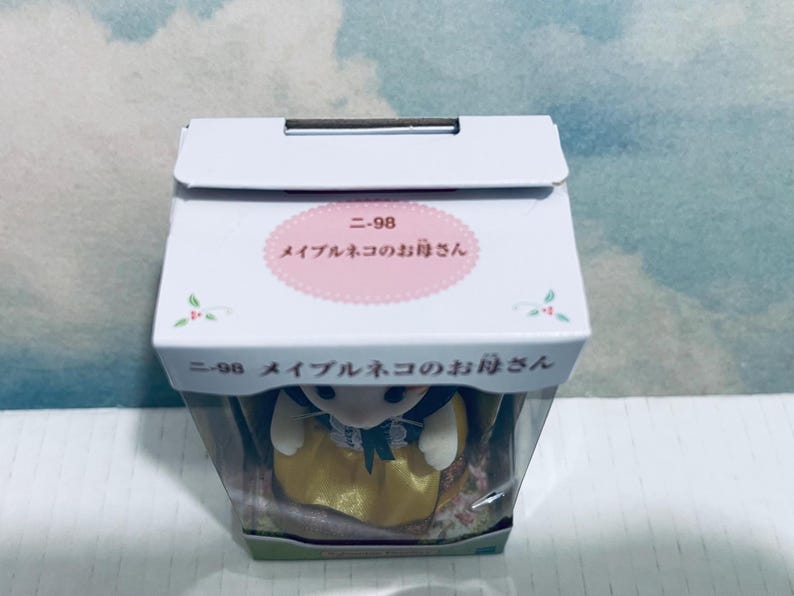 Pu&ograve; includere: Una scatola bianca con testo giapponese e un'etichetta ovale rosa. L'etichetta recita "ニ-98 メイプルネコのお母さん". All'interno della scatola c'&egrave; una piccola statuina di un gatto con un vestito giallo e un grembiule bianco.