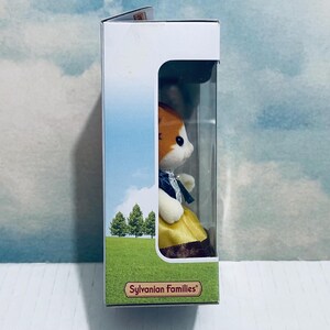 Pu&ograve; includere: Figurina Sylvanian Families di un gatto con abito giallo e fiocco blu, esposta in una scatola con finestra trasparente. La scatola presenta un paesaggio con alberi e il nome del marchio.