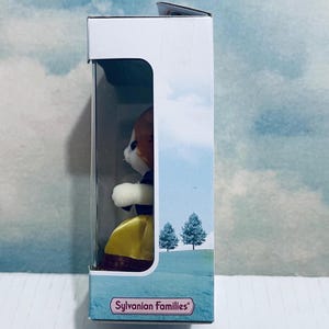 Pu&ograve; includere: Una figura giocattolo Sylvanian Families di una volpe in una scatola. La volpe ha pelliccia marrone e bianca, indossa un vestito giallo e un colletto viola. La scatola presenta una scena paesaggistica con due alberi e il logo Sylvanian Families.