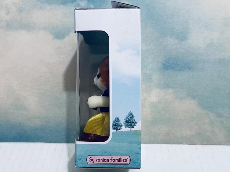 Pu&ograve; includere: Una figura giocattolo Sylvanian Families di una volpe in una scatola. La volpe ha pelliccia marrone e bianca, indossa un vestito giallo e un colletto viola. La scatola presenta una scena paesaggistica con due alberi e il logo Sylvanian Families.