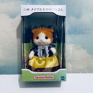 Pu&ograve; includere: Una figura di gatto giocattolo Sylvanian Families in una scatola. Il gatto ha pelliccia arancione e bianca e indossa un vestito blu scuro con una gonna gialla. La scatola ha testo giapponese e il logo Sylvanian Families.