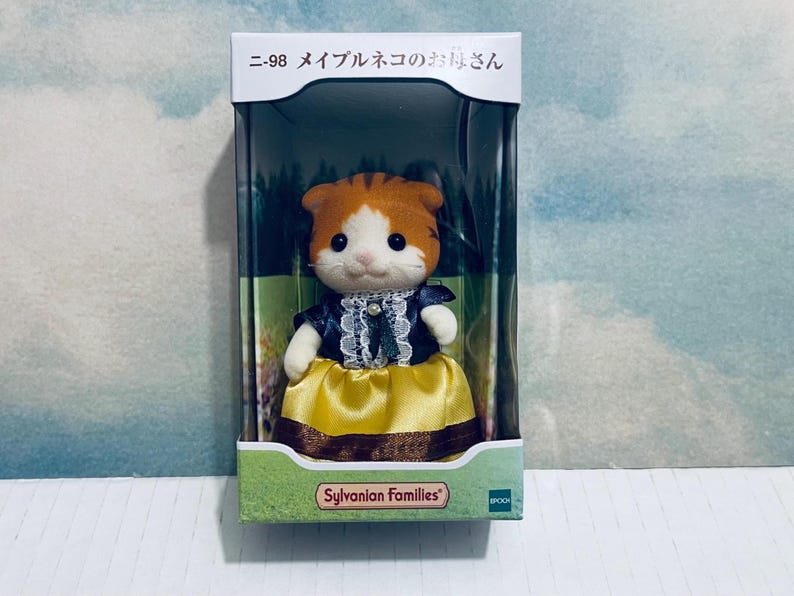 Pu&ograve; includere: Una figura di gatto giocattolo Sylvanian Families in una scatola. Il gatto ha pelliccia arancione e bianca e indossa un vestito blu scuro con una gonna gialla. La scatola ha testo giapponese e il logo Sylvanian Families.