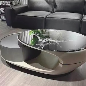 May include: Contemporary oval coffee table with a black marble-effect top and a grey and white base. The table features a distinctive design with a lower shelf, complemented by a black leather sofa. The room has a modern aesthetic.