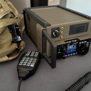May include: A tan and black radio communication device with a microphone and a backpack. The radio has a digital display and various knobs and buttons. The microphone is black with multiple buttons. The backpack is tan.