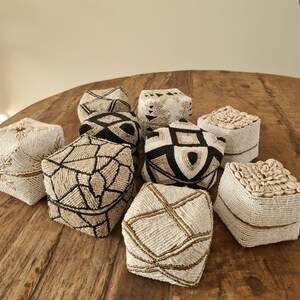 Op de afbeelding: Een verzameling kralenboxen met geometrische patronen in zwart, wit en beige. Sommige dozen zijn versierd met schelpen. De dozen zijn gerangschikt op een houten oppervlak, waarbij hun ingewikkelde ontwerpen en texturen worden getoond.