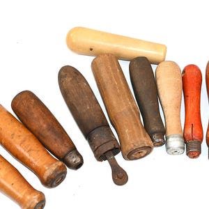May include: A collection of nine assorted tool handles made from various types of wood, including light, medium, and dark brown tones, with one handle painted bright orange and one smooth cream-colored handle, featuring metal ferrules.